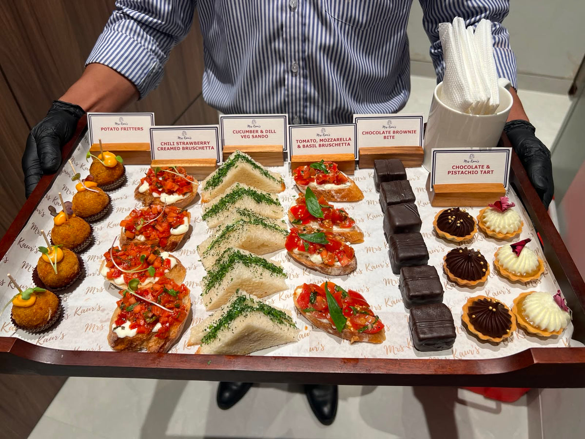 A Mrs Kaur’s server holding a tray of catering miniatures: potato fritters, bruschetta, cucumber and dill sandos, brownie bites and tarts