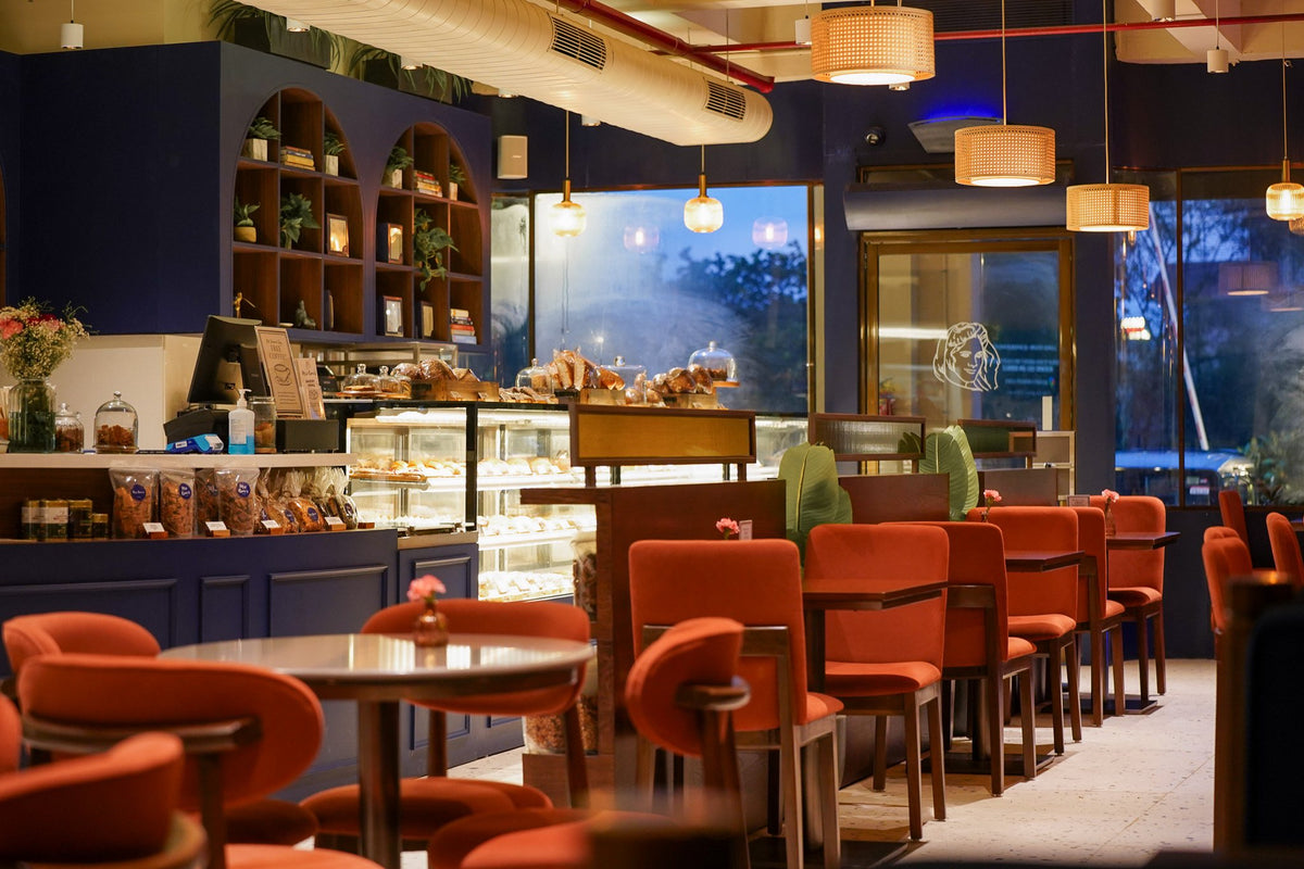The café at Mrs Kaur’s on Golf Course Road, Gurgaon, in the evening: the bakery counter, terracotta chairs, and the drawn portrait of Pritam Kaur on the glass door