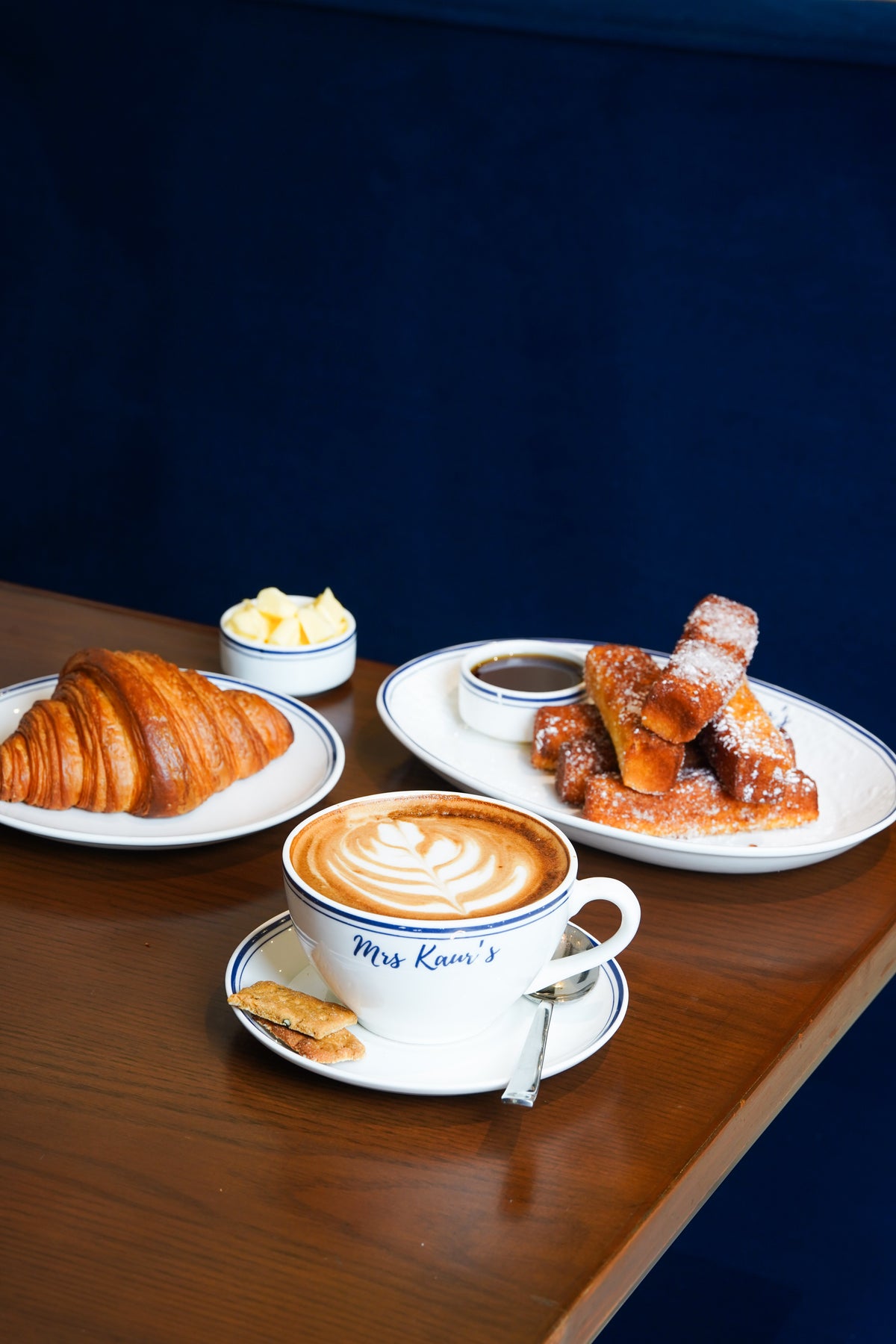 A latte in a Mrs Kaur’s cup with a croissant and French toast at a café table